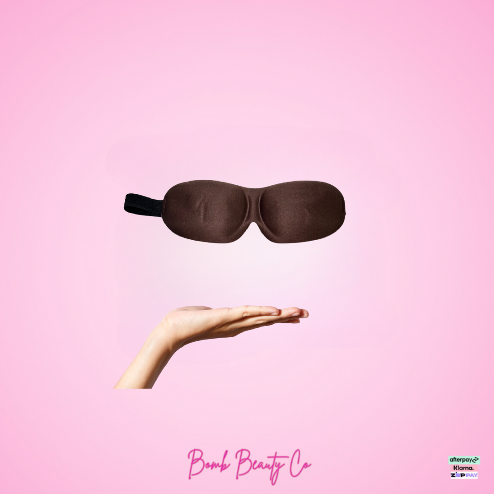 Brown sleep mask held by a hand on a pink background with 'Bomb Beauty Co' branding.
